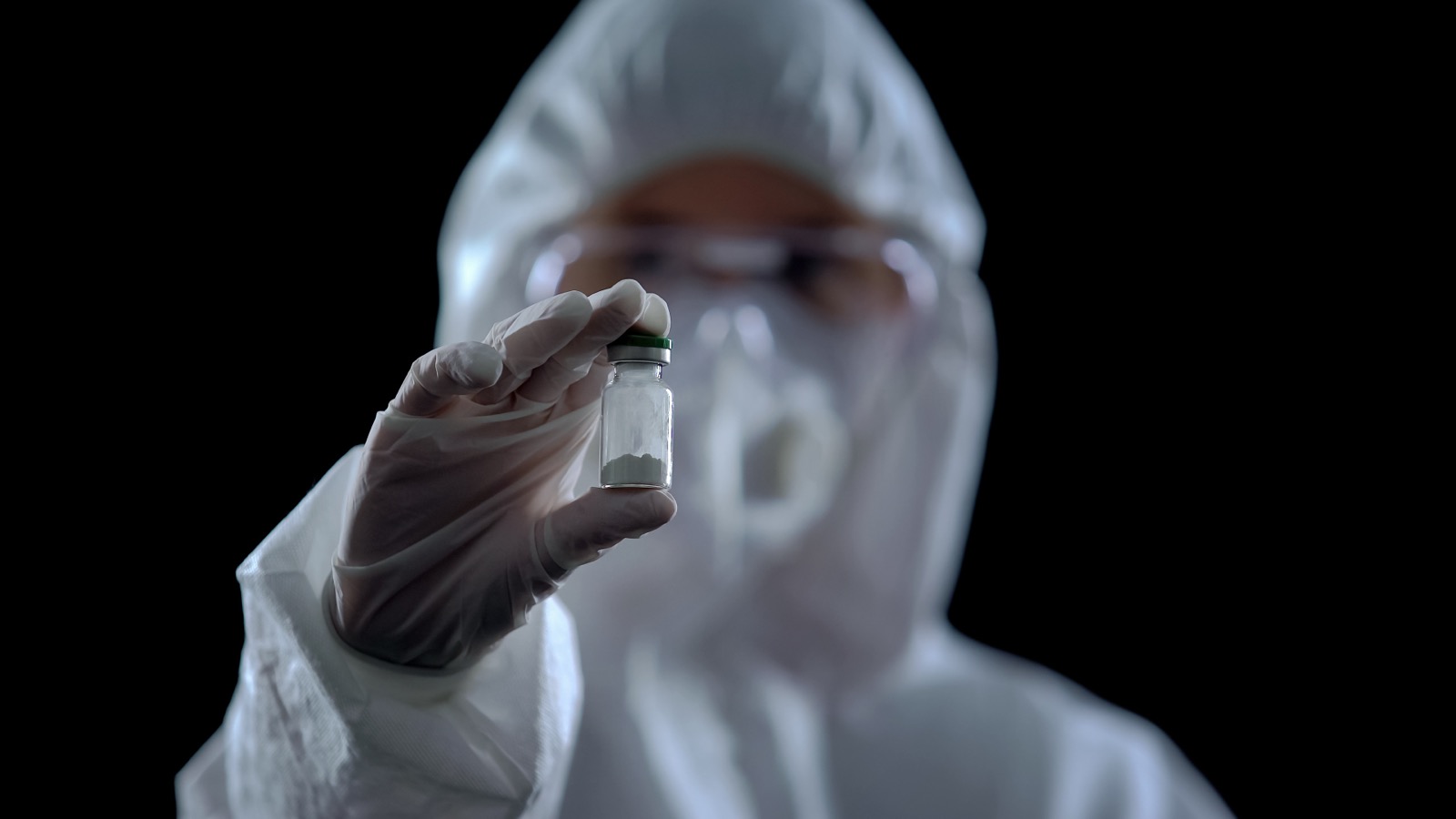 Person in a full hazmat suit holding a small pharmaceutical vial against a dark background